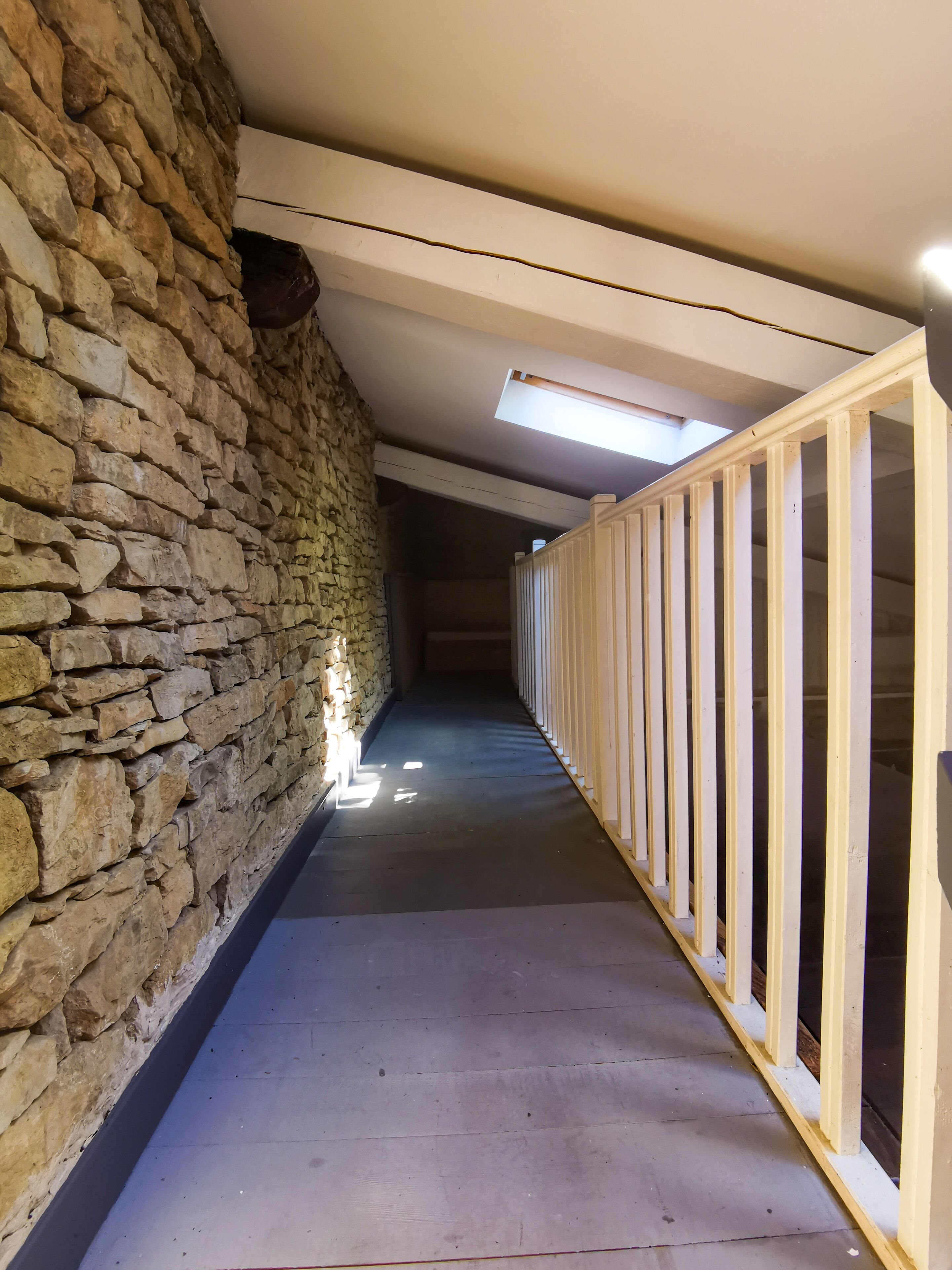 Un couloir aux murs de pierre, aux rampes en bois et doté d’une verrière.
