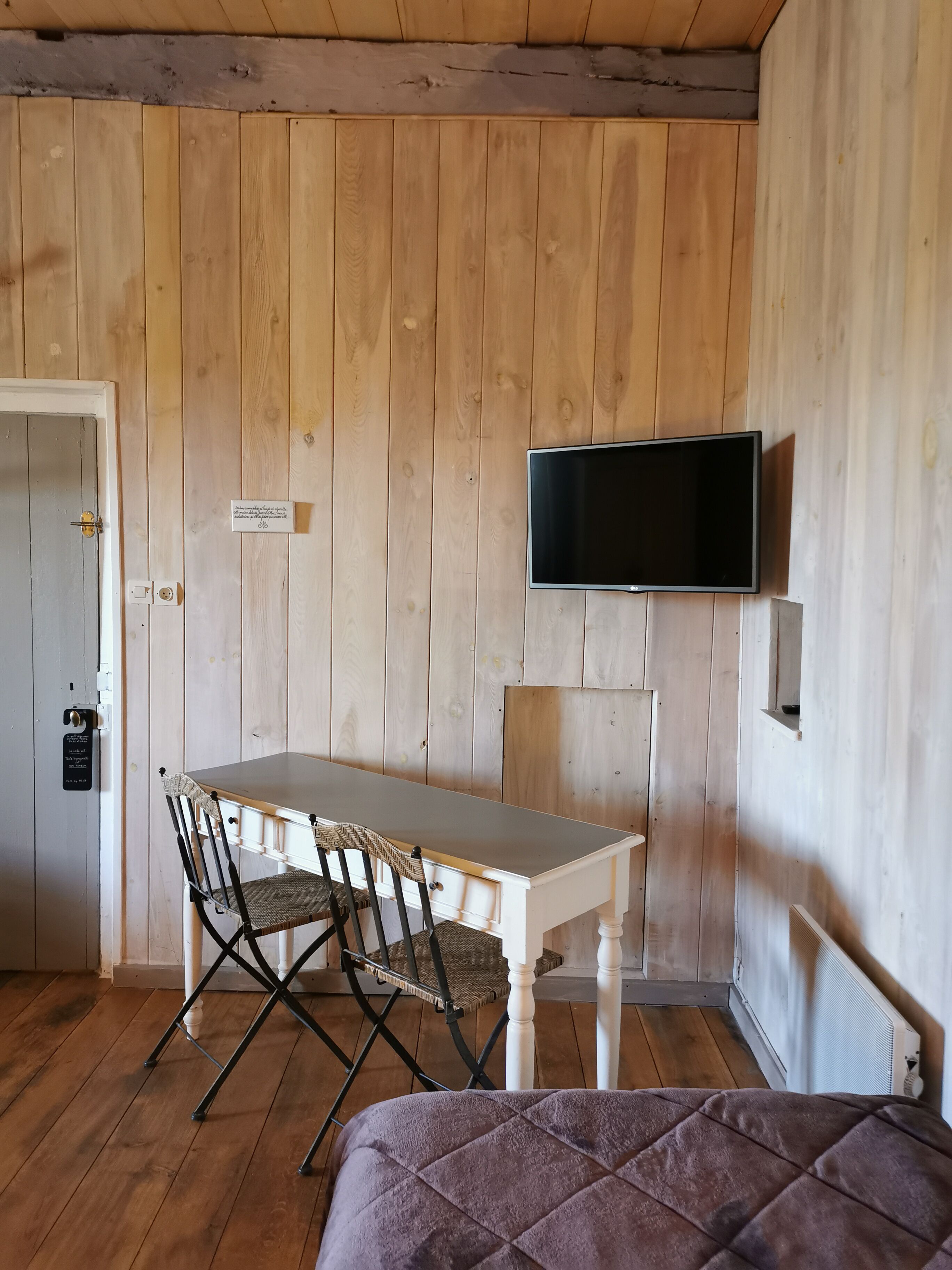 Una habitación con paredes de madera, una mesa blanca con sillas, un televisor de pantalla plana en la pared y una cama con una manta morada.