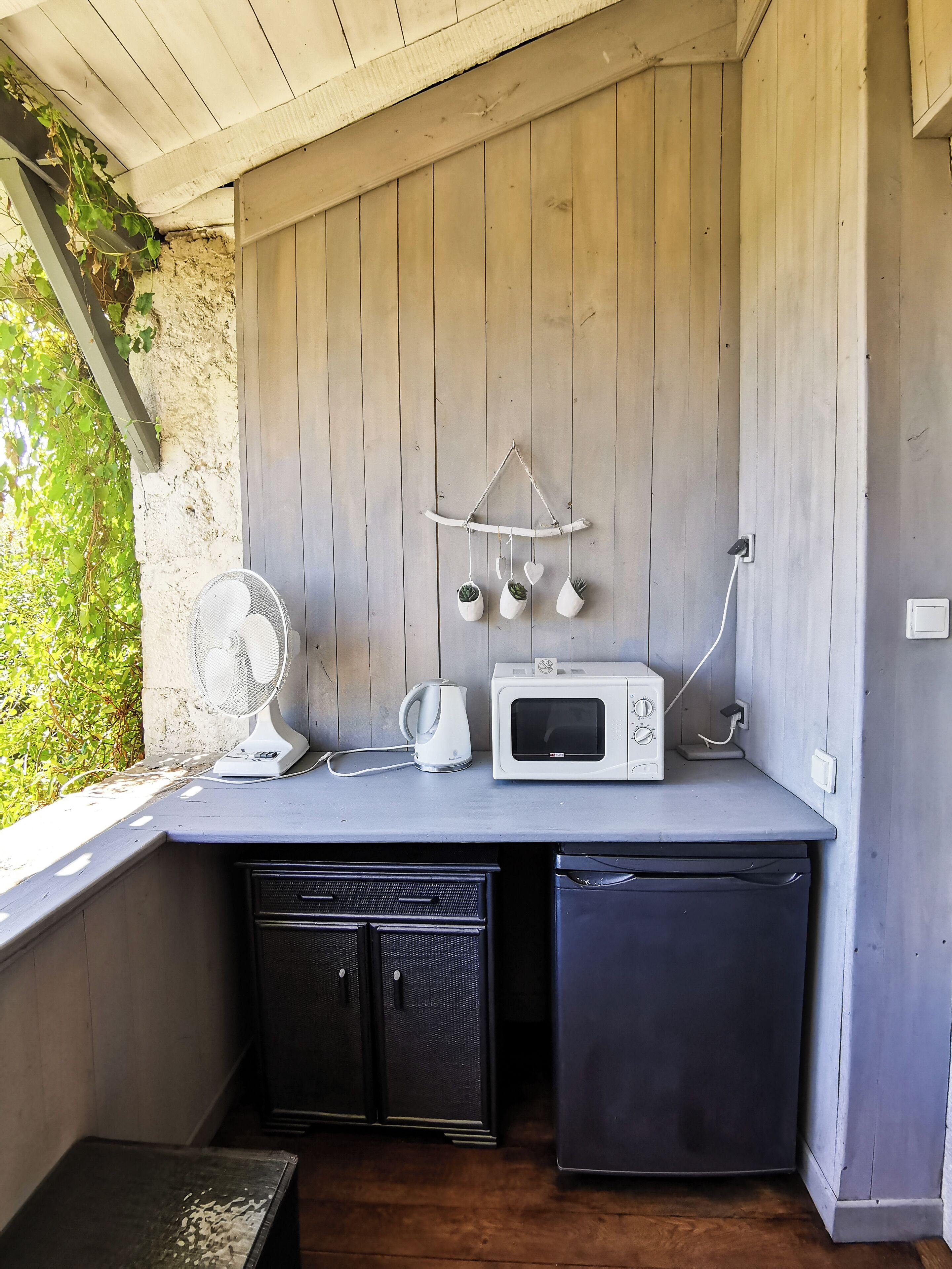 Una cocina compacta con una pared de madera, un ventilador blanco, una tetera blanca y un microondas blanco.