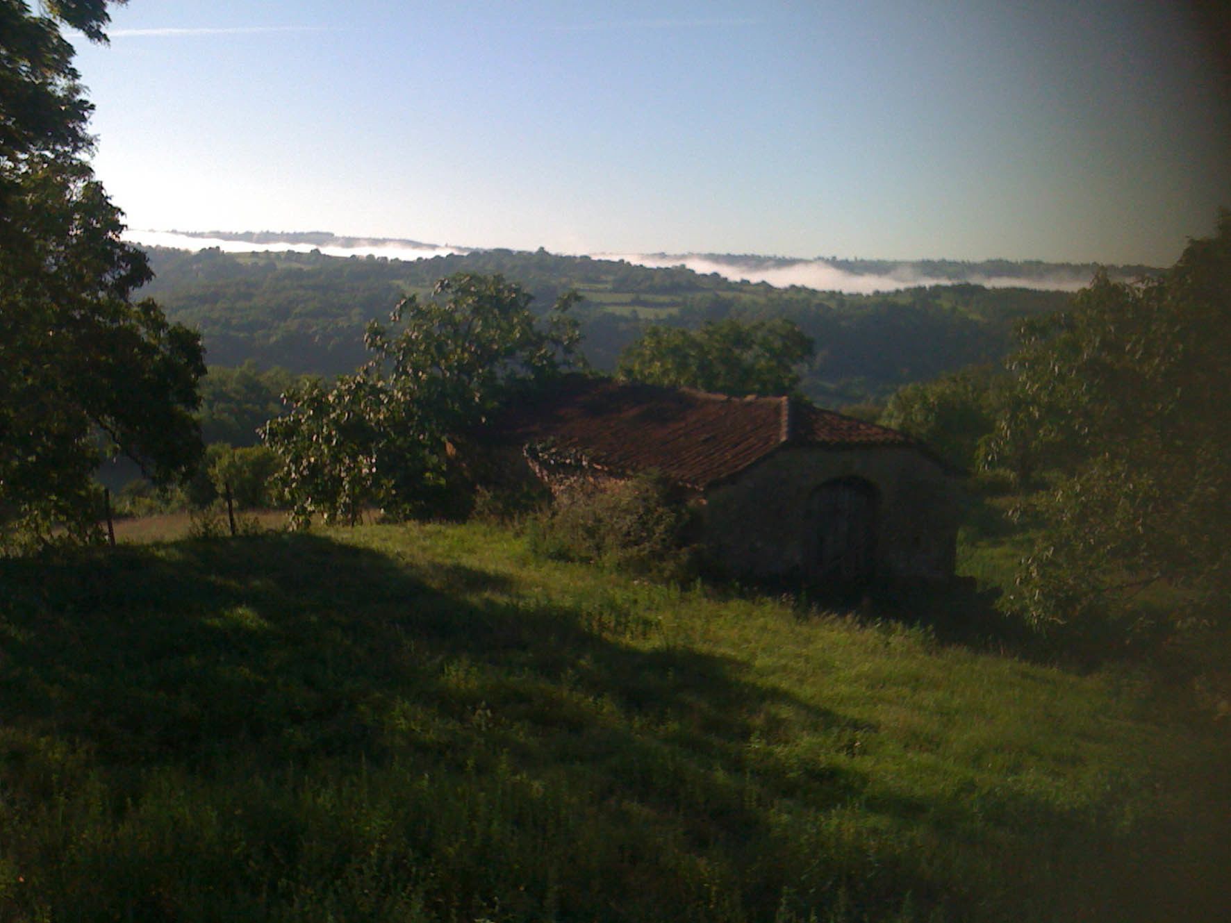 A rural landscape with a small stone building, surrounded by trees and fields.