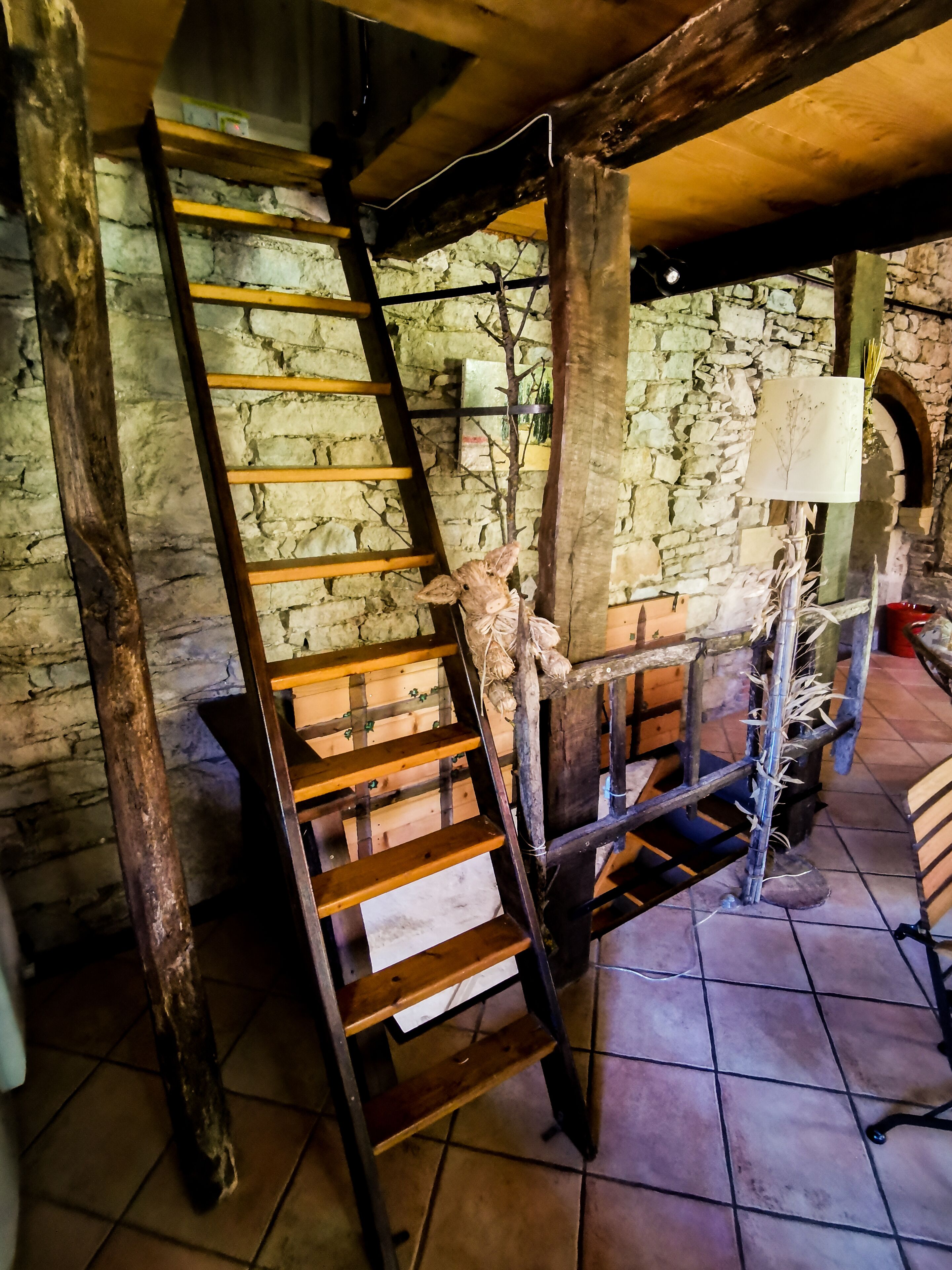 Un escalier rustique avec des marches en bois et une rampe en métal, menant à un mur de pierre surmonté d’une porte en bois.