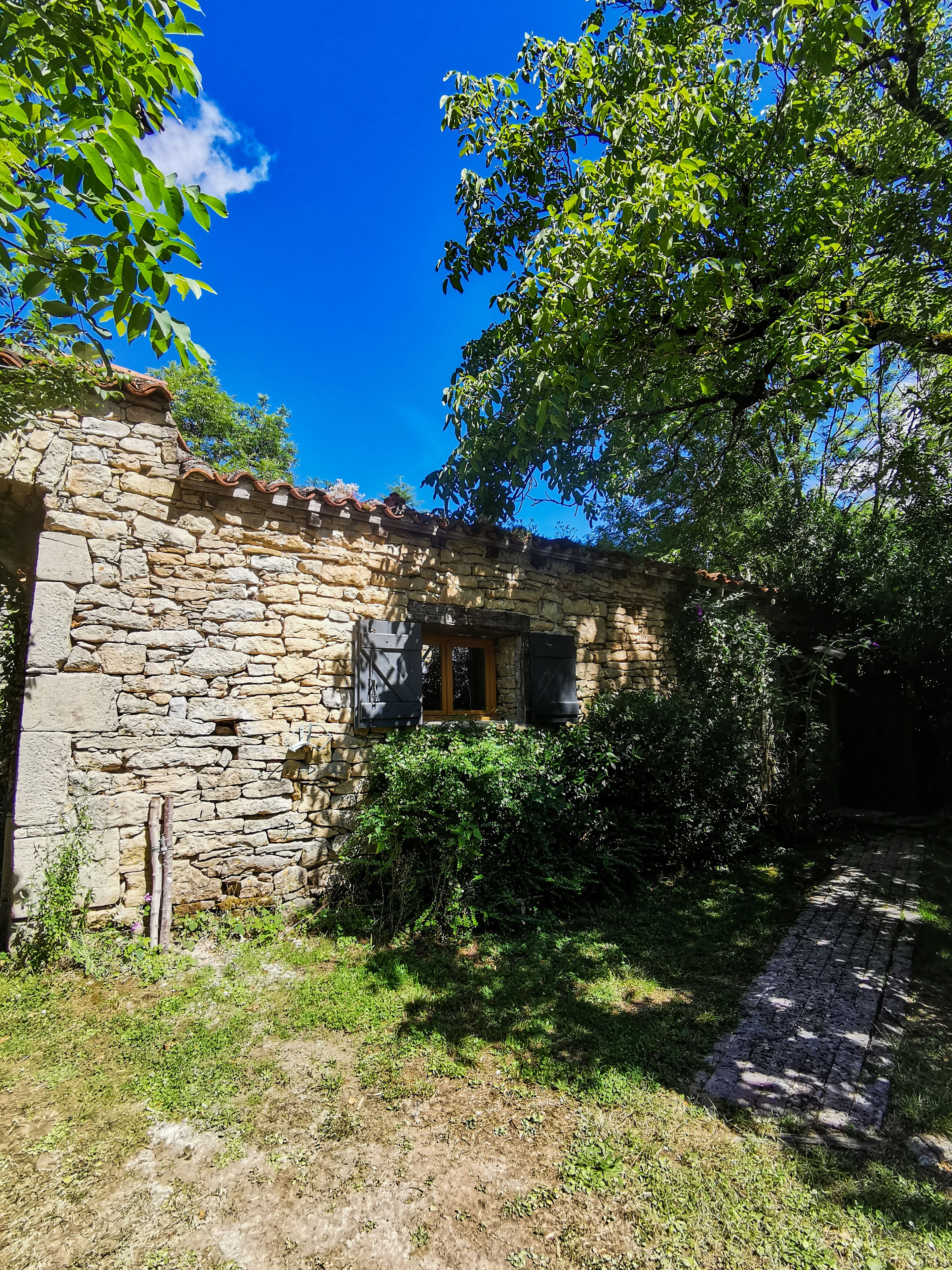 A stone building with a wooden door and window shutters, surrounded by greenery and a clear blue sky.