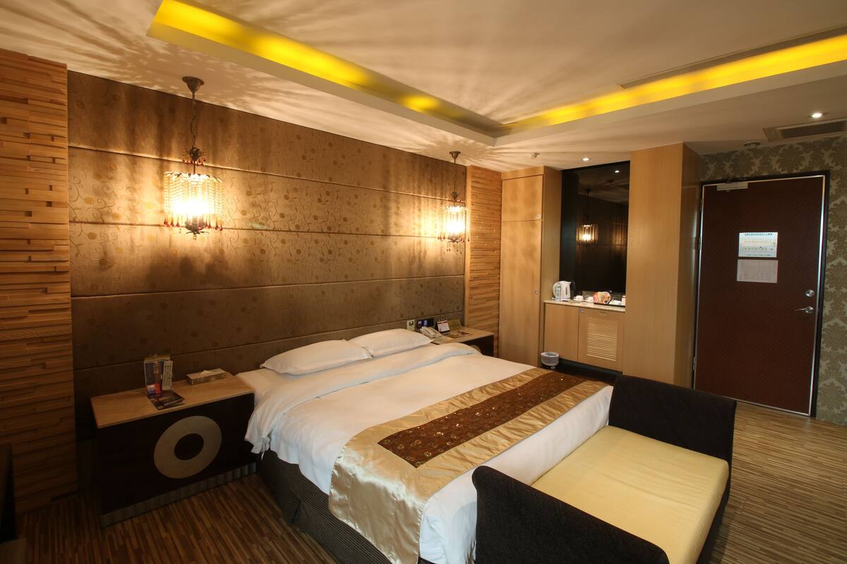 Une chambre d’hôtel avec un grand lit, une tête de lit en bois, deux lampes fixées au mur, une table de chevet avec un téléphone et un livre, et une porte dotée d’un miroir.