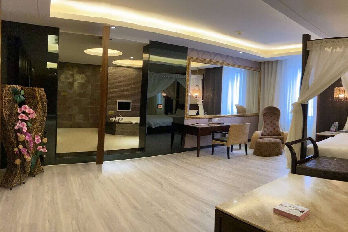Une chambre d’hôtel comprenant un lit, une table de chevet, une chaise, un canapé, un miroir et une salle de bain avec une baignoire et un lavabo.