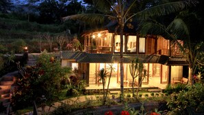 Una casa de dos pisos con un balcón, rodeada de vegetación tropical y una escalera de piedra.