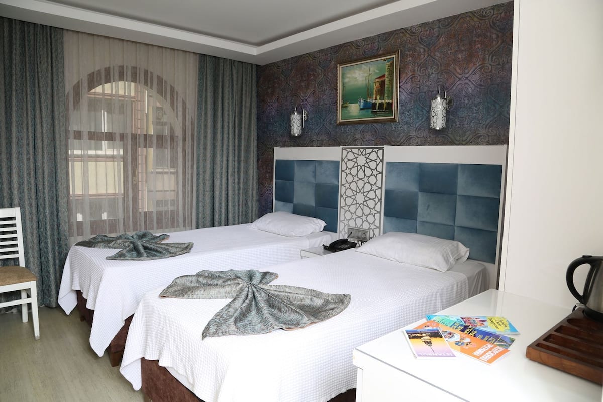 Une chambre d’hôtel avec deux lits, un bureau, une chaise, une fenêtre avec des rideaux, un mur décoratif et un tableau.