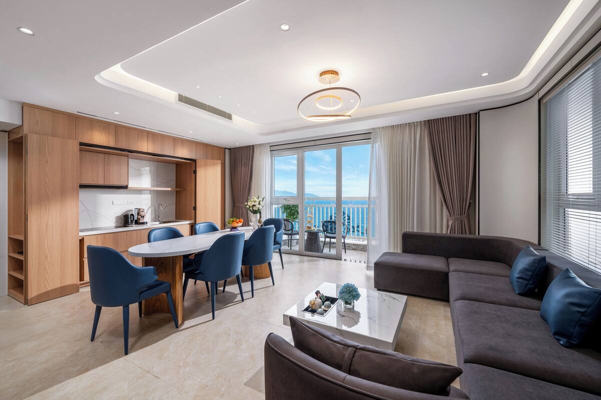 A modern living room with a sofa and coffee table, a dining area with chairs and a table, and a kitchen with wooden cabinets and a window overlooking a balcony.