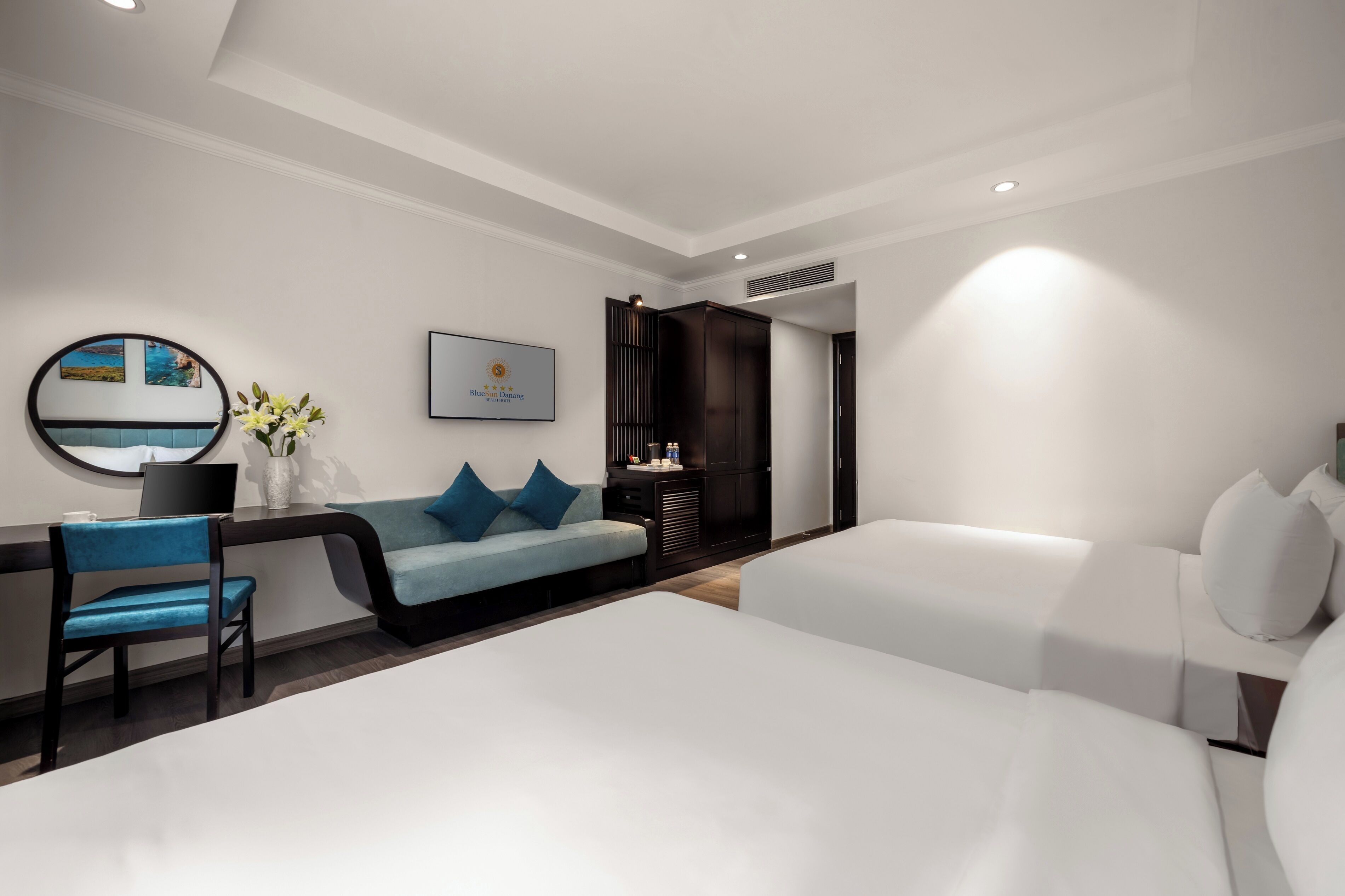 A modern hotel room with a large bed, a desk with a chair, a sofa, and a small table with a vase of flowers.