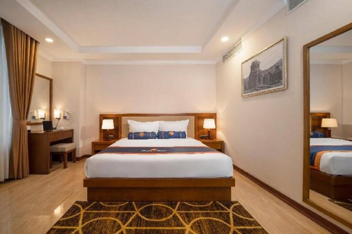 A hotel room with a large bed, bedside tables with lamps, a desk, a chair, a patterned rug, and a framed picture on the wall.