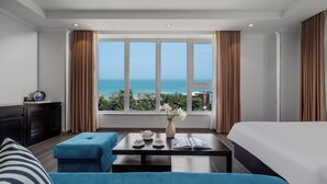 Un cuarto de hotel con un ventanal grande, una cama, un sofá azul, una mesita con un florero y vista al océano.