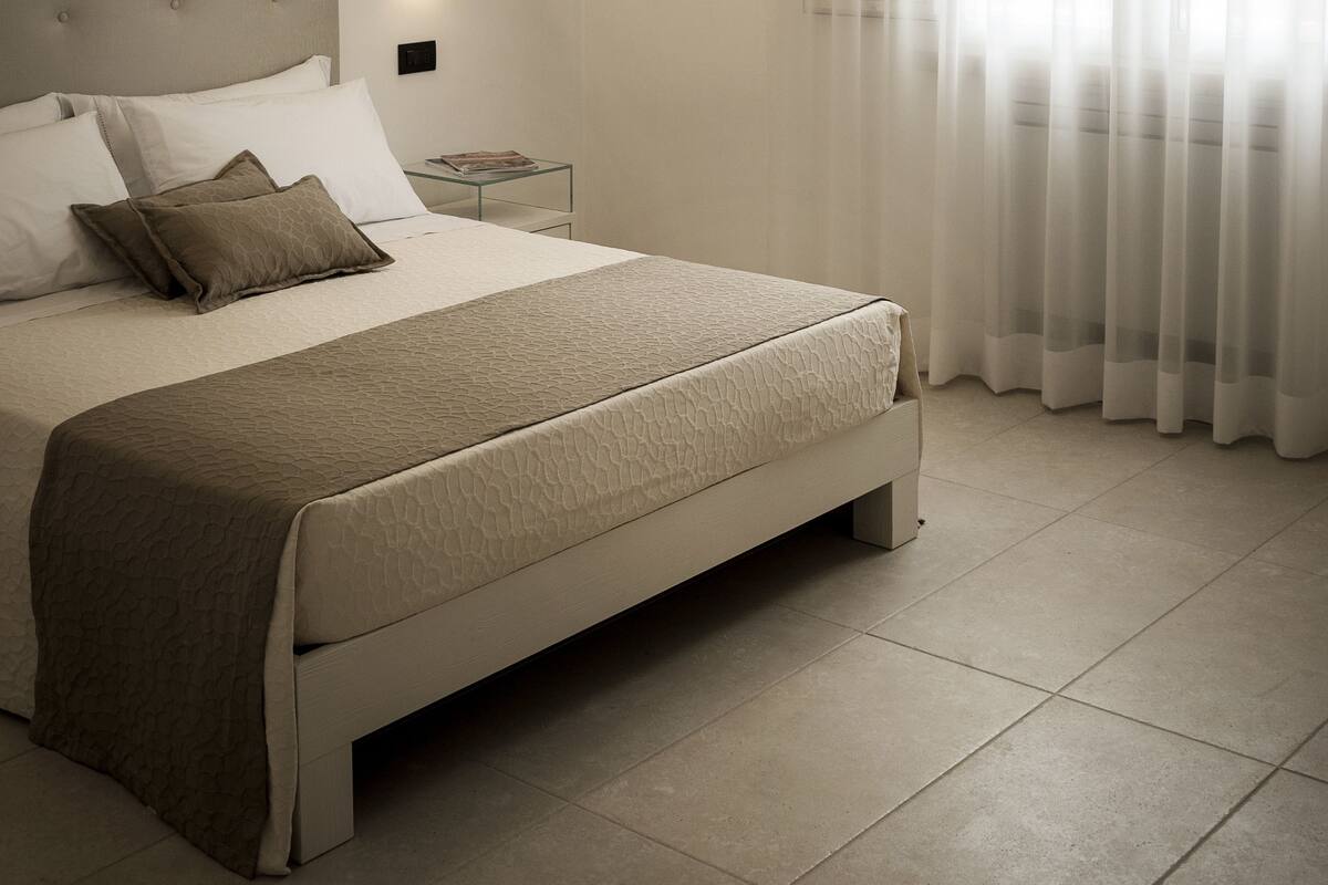 A modern bedroom with a large bed, a headboard, a bedside table, and a window with sheer curtains.