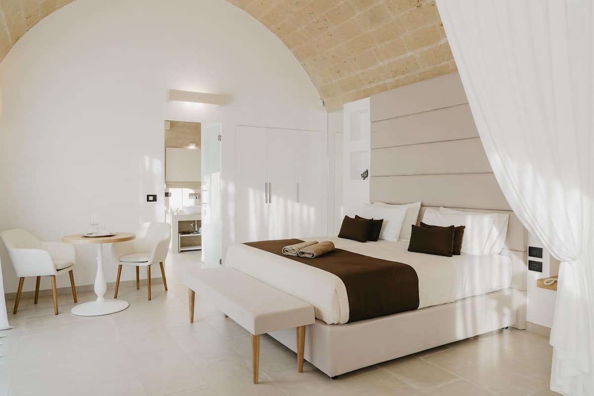 A modern bedroom with a large bed, a bedside table, a bench, and a small table with chairs.