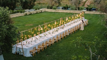 An outdoor dining area with a long table set for a meal, surrounded by chairs and decorated with string lights and greenery.