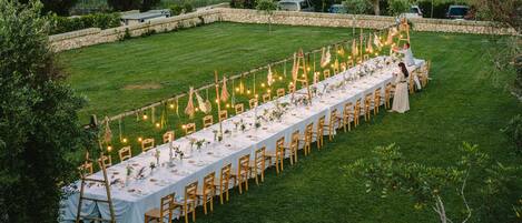 Un espace de restauration en plein air avec une longue table dressée pour un repas, entourée de chaises et décorée de guirlandes lumineuses et de verdure.