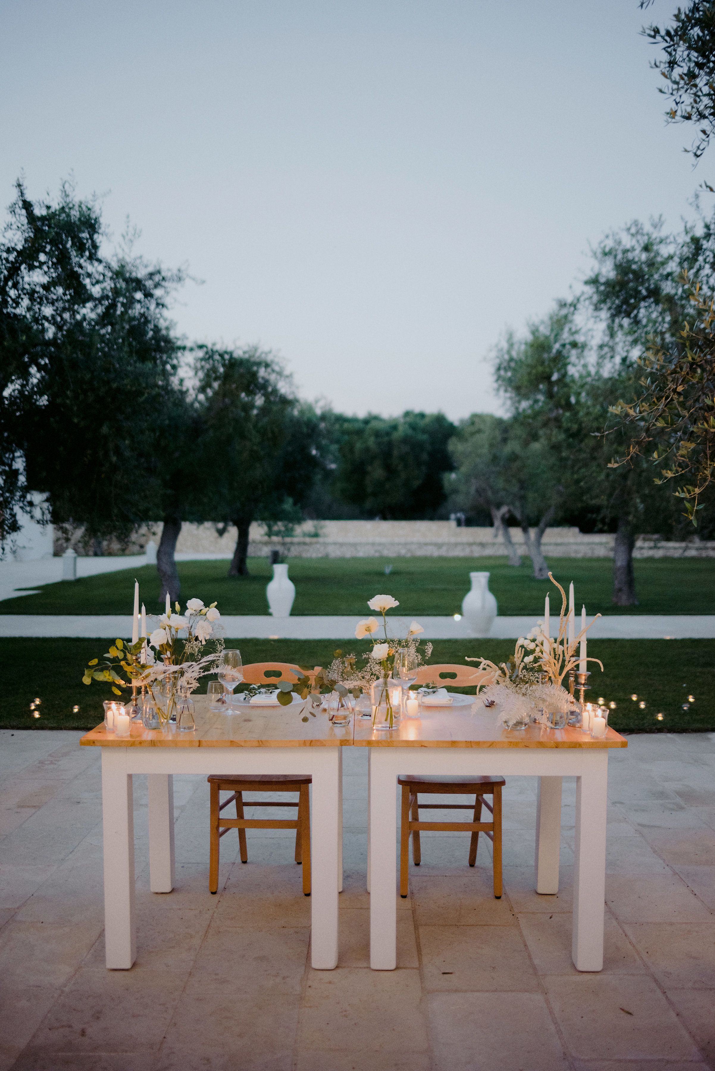 Una mesa de comedor al aire libre con velas y flores, rodeada de árboles y un florero blanco en un jardín.