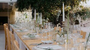 Una larga mesa de madera dispuesta para un evento al aire libre elegante, con manteles blancos, velas y cristalería.