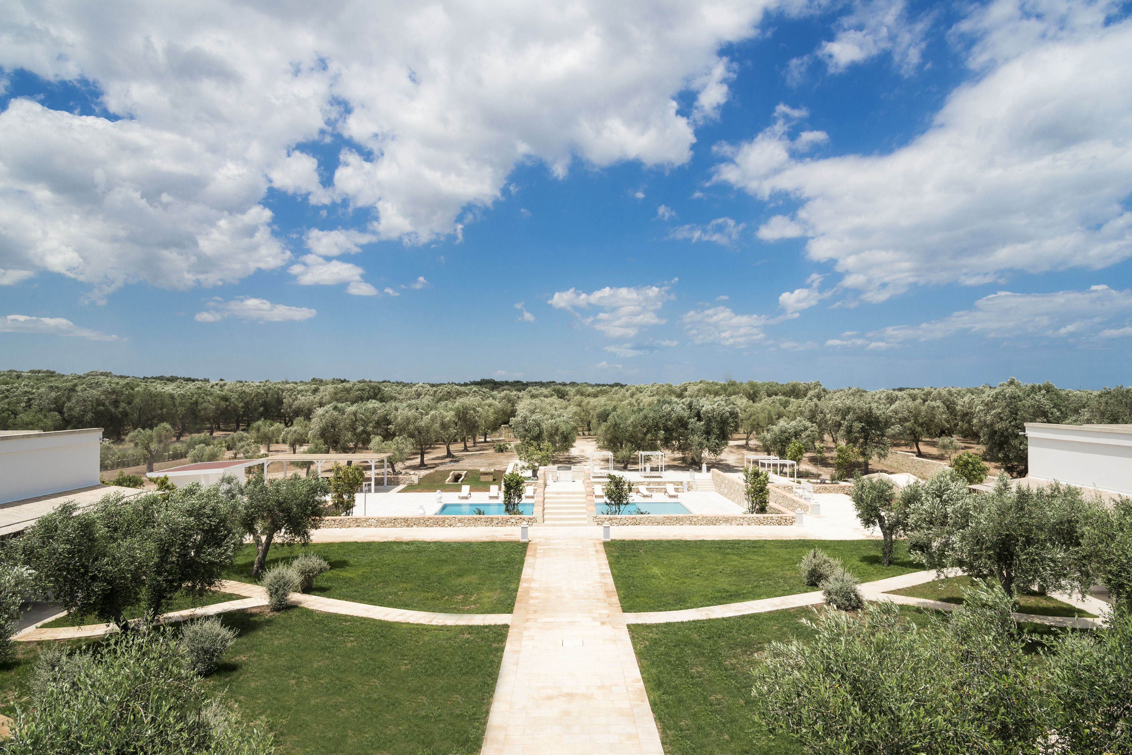 Un complejo turístico con un camino central que lleva a una zona de piscina, rodeado de olivos y un cielo azul despejado.