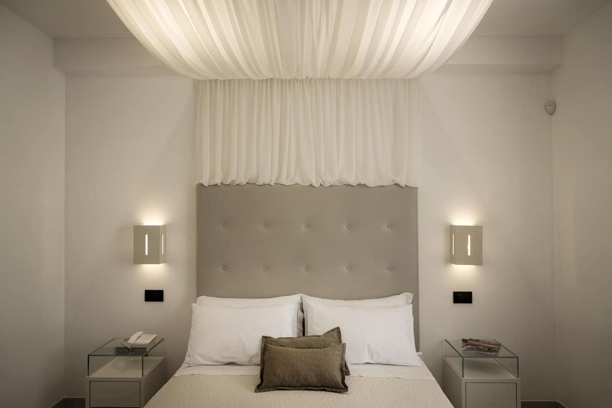 A bedroom with a tufted headboard, two bedside tables with lamps, a bed with white linens, and a sheer white drape over the window.