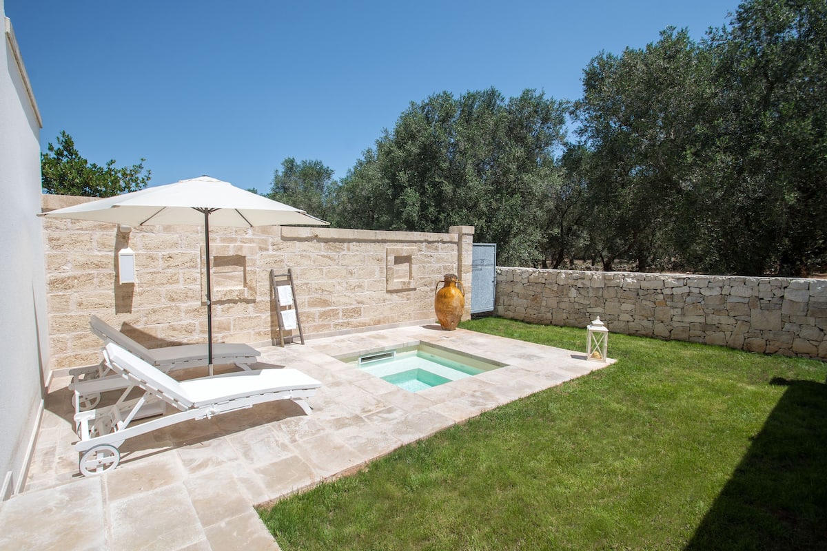A backyard with a swimming pool, a lounge chair, a sun umbrella, and a stone wall.