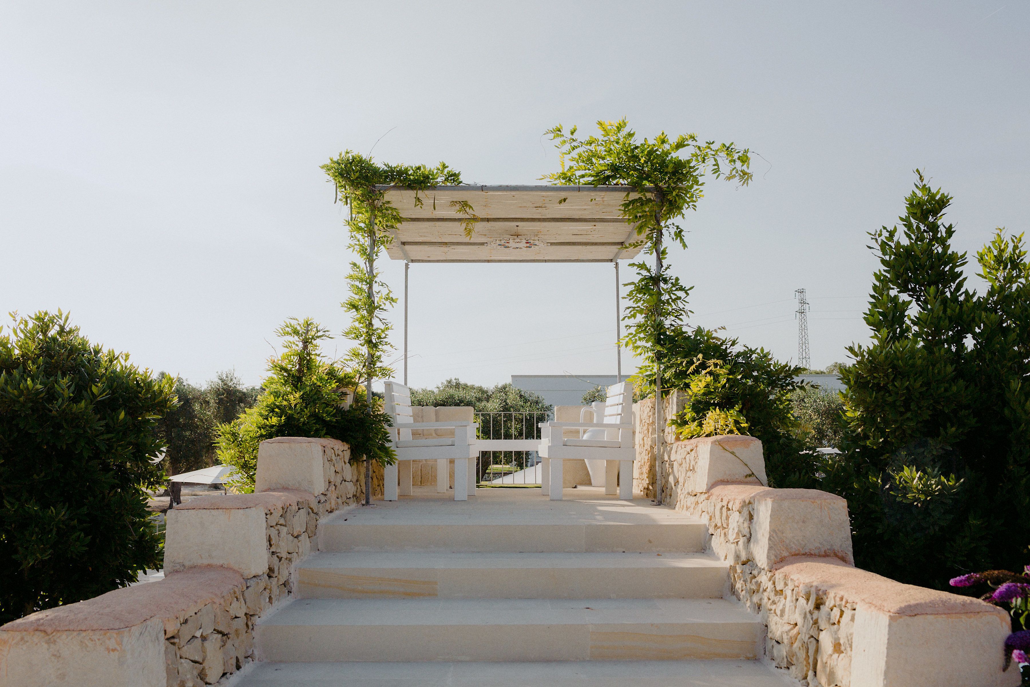 Una scala in pietra conduce a un arco in legno bianco, adornato da vegetazione.