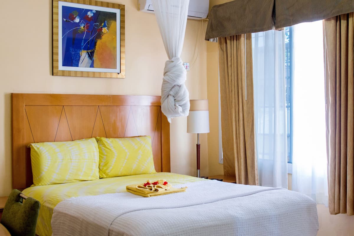 A hotel room with a wooden headboard, a bed with yellow and white bedding, a painting on the wall, a bedside lamp, and a window with curtains.