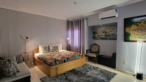 A bedroom with a wooden bed, a chair, a bedside table, a large painting, and an air conditioning unit.
