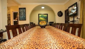 A long dining table with a leopard print tablecloth, surrounded by wooden chairs. There is a painting on the wall and a television mounted on the wall. - Alma House (Accra)