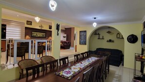 A dining area with a long wooden table, chairs, and a patterned table runner. There is a sofa and a television in the background.
