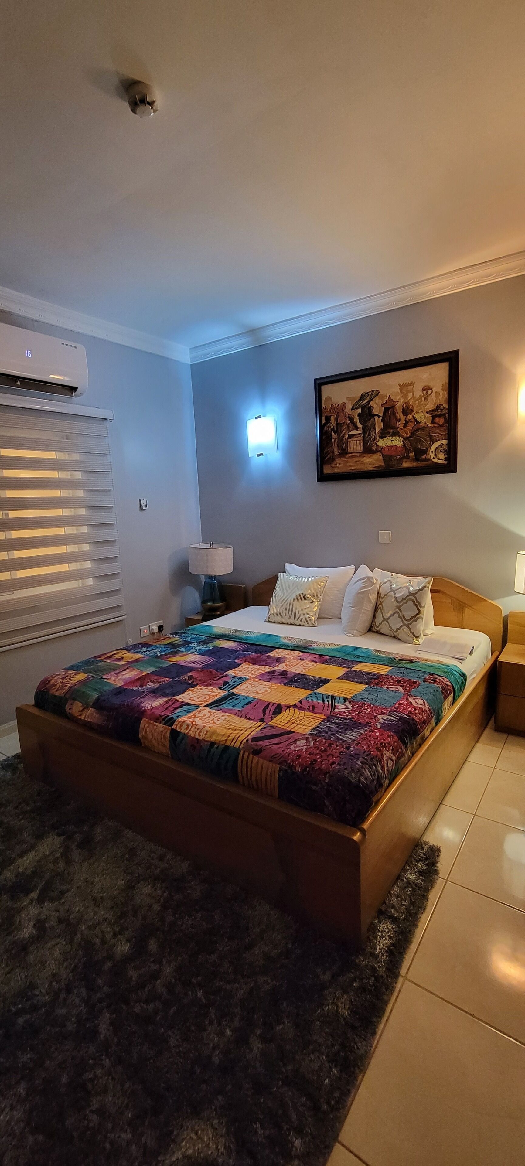 A bedroom with a wooden bed, a colorful quilt, a wall-mounted air conditioner, a window with blinds, and a framed picture on the wall.