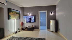 A living room with a flat-screen TV mounted on the wall, a sofa, a coffee table, a refrigerator, and a door leading to another room.