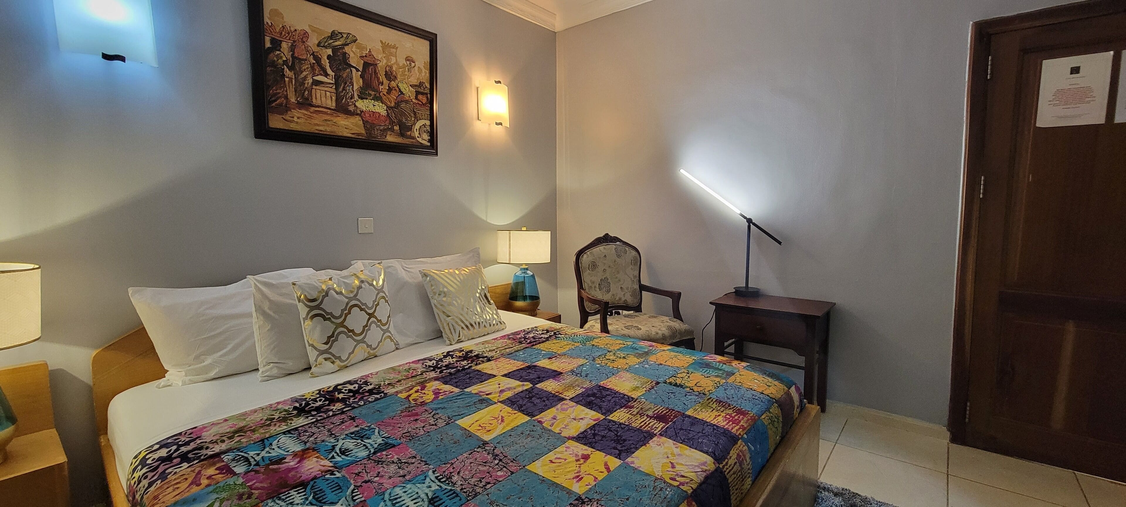 Un dormitorio con cama, una silla, una mesita de noche, una lámpara y un cuadro en la pared.