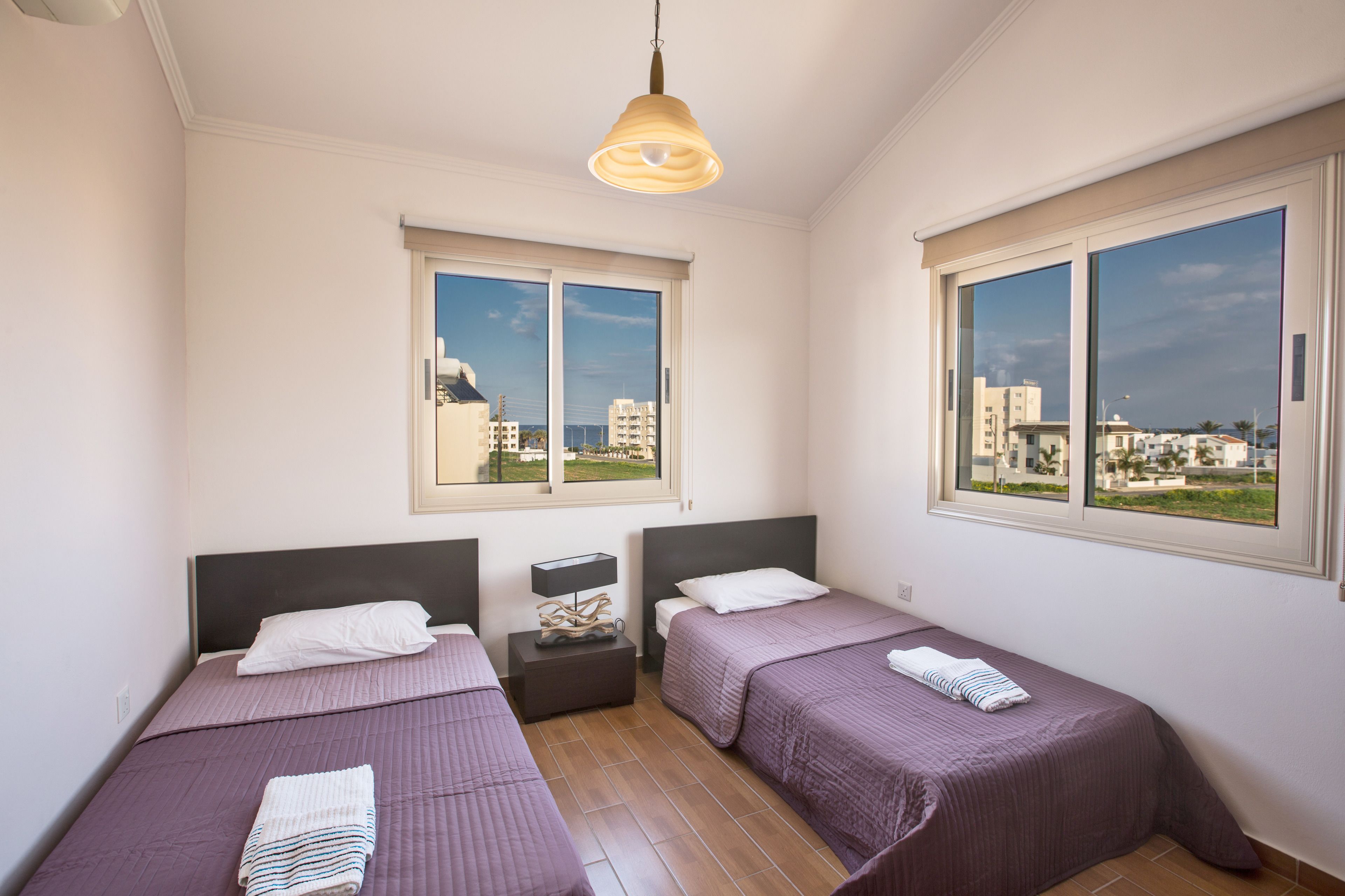 A hotel room with two beds, a nightstand, a lamp, and large windows offering a city view.