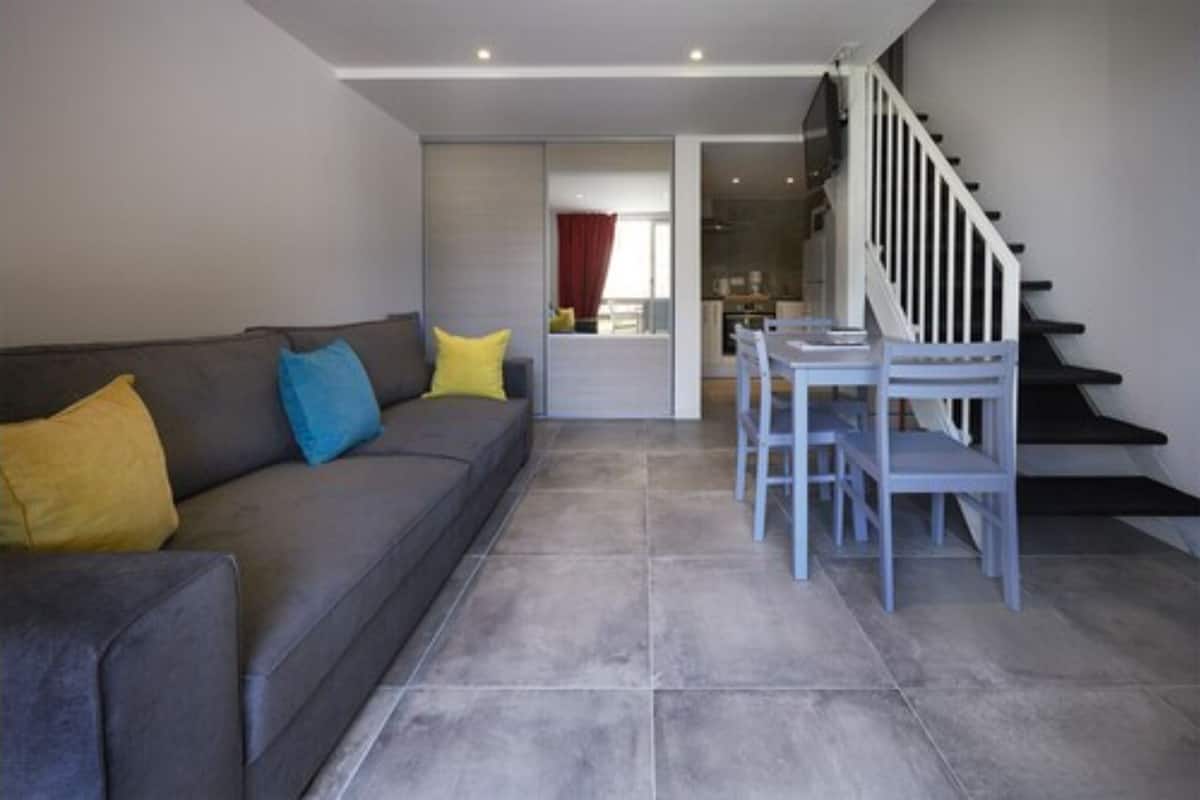 Una sala de estar con un sofá gris, cojines azules y amarillos, una mesa de comedor con sillas blancas y una escalera.