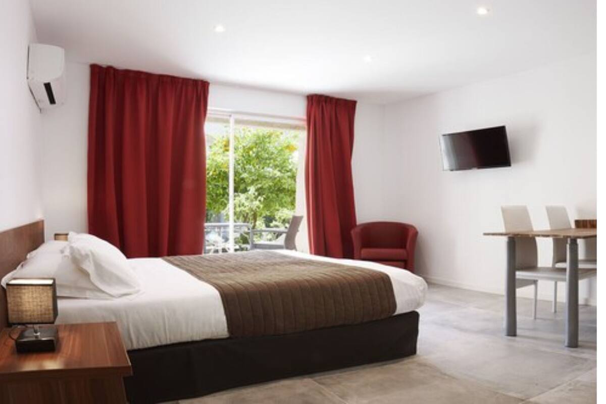 Una habitación de hotel moderna con cama, escritorio, silla, televisor y balcón con cortinas rojas.