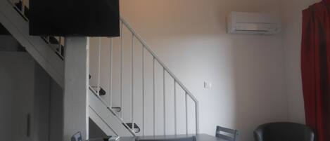 A room with a dining table and chairs, a staircase, a TV mounted on the wall, and an air conditioning unit.