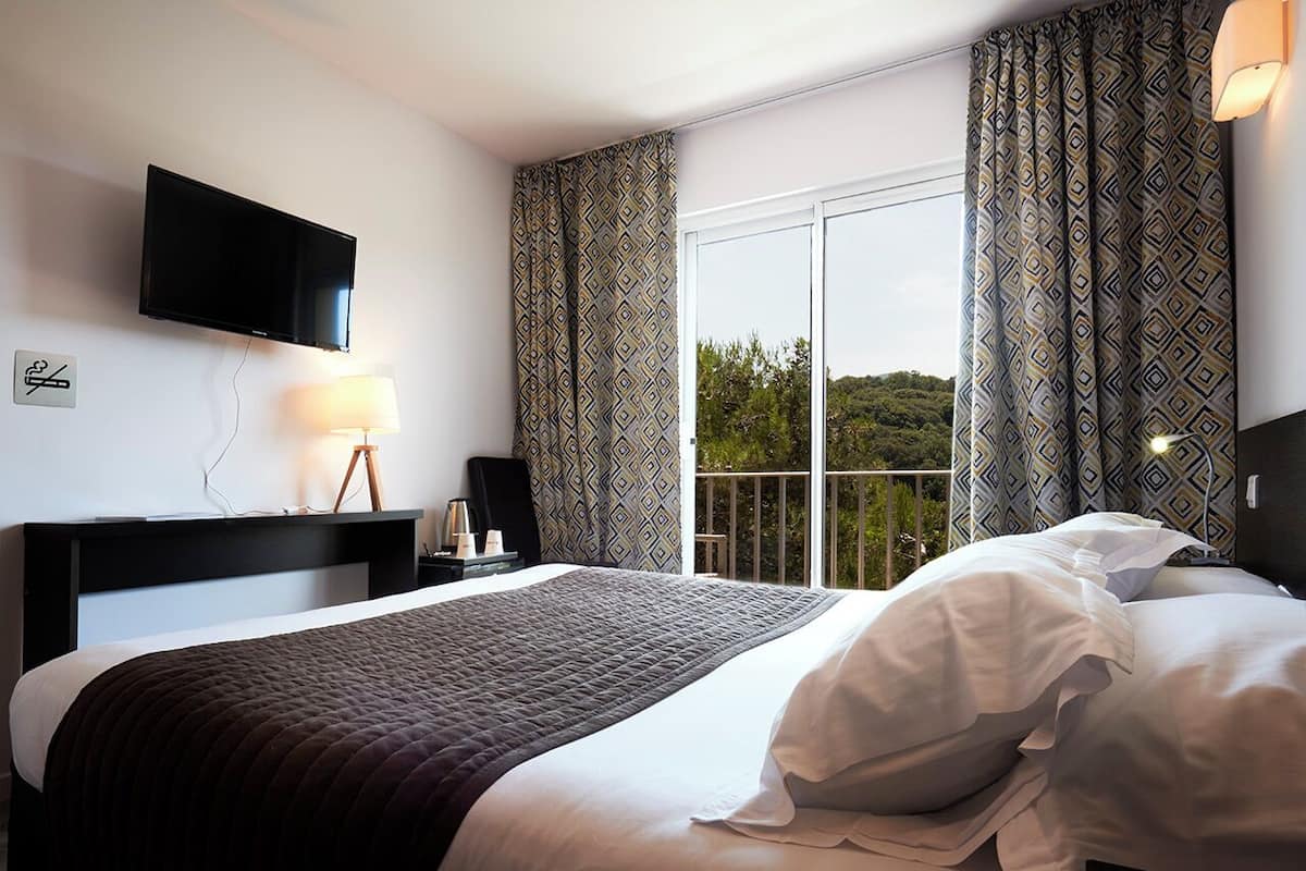 Habitación de hotel con cama, televisor montado en la pared, ventana con cortinas y mesita de noche con lámpara.