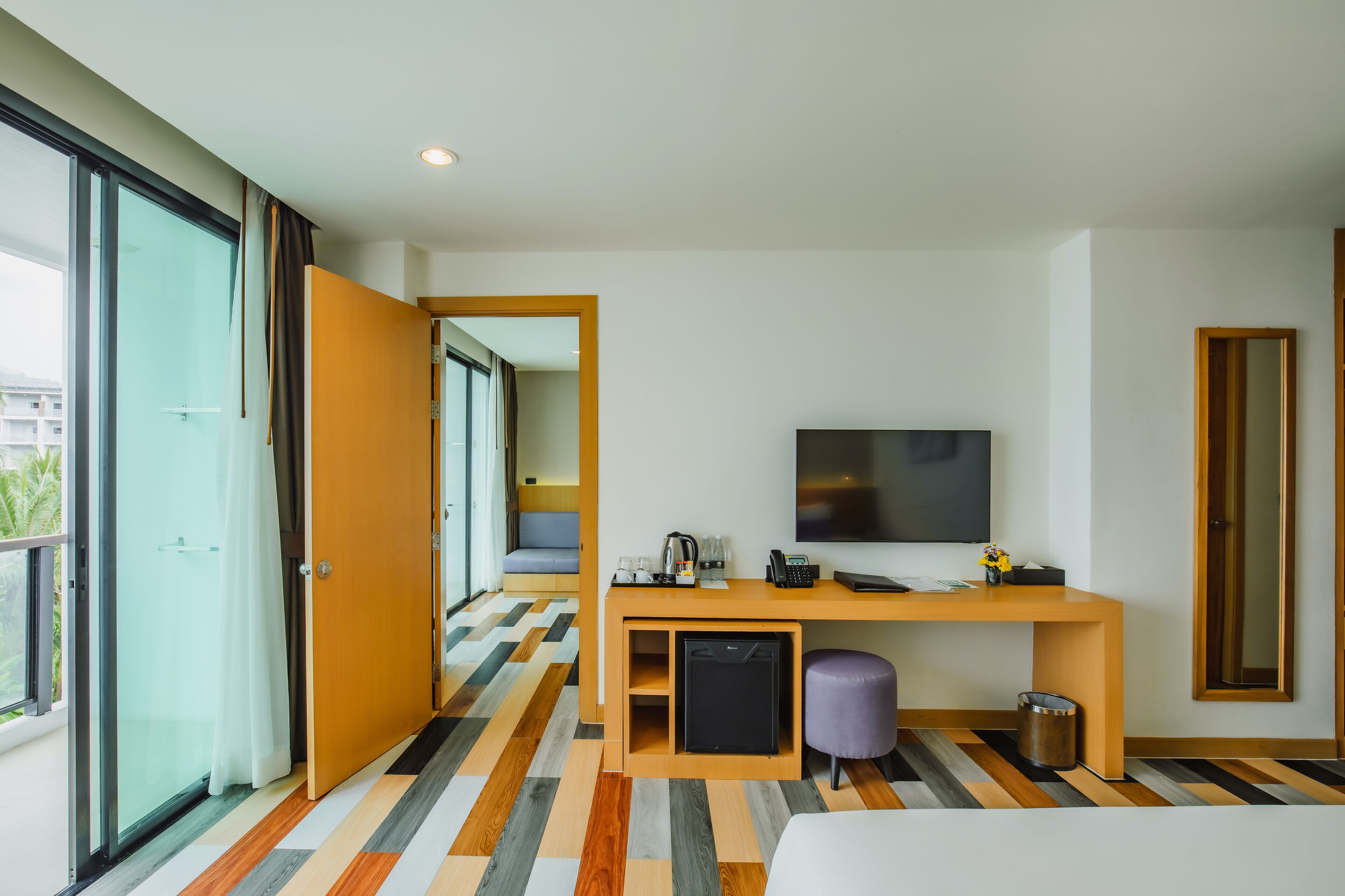 A modern hotel room with a large window, a flat-screen TV, a desk with a coffee maker, and a compact refrigerator.