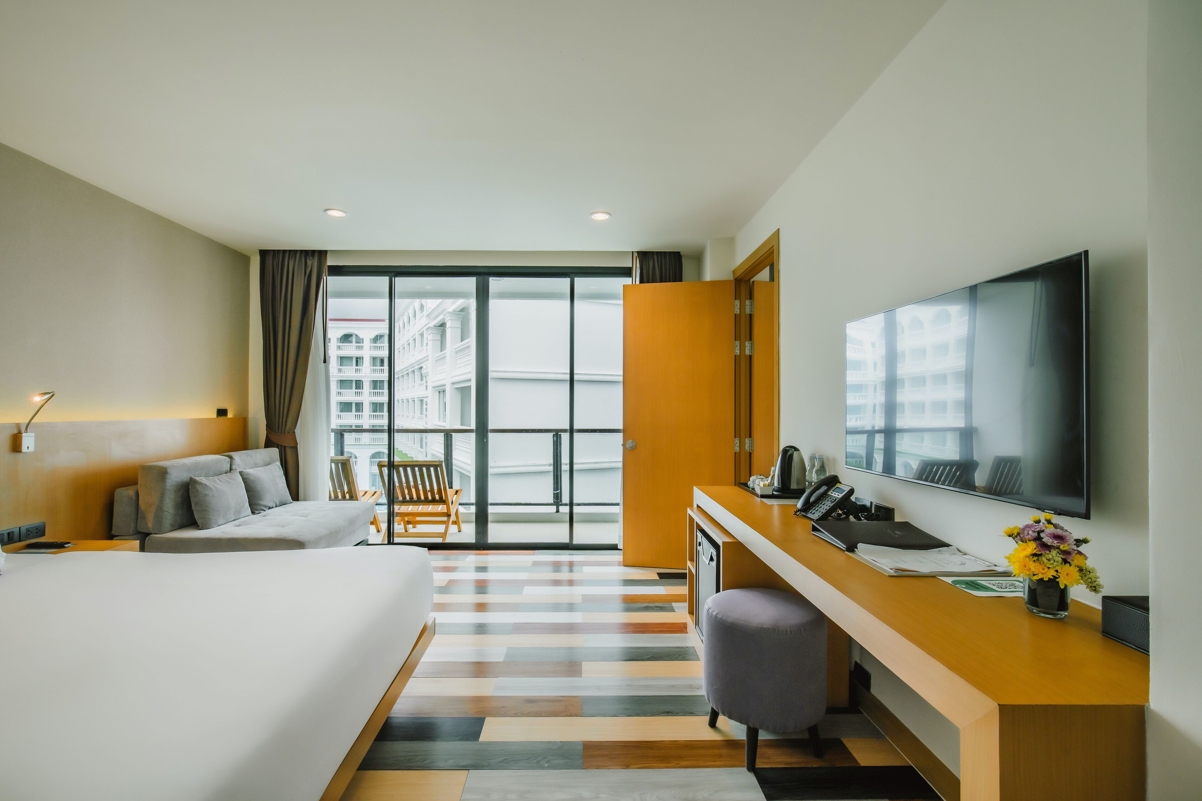 A modern hotel room with a large bed, a desk with a computer, a flat-screen TV, and a balcony with a city view.