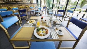 A dining area with tables and chairs, a breakfast setting with plates, eggs, and beverages, and a view through large windows. - The Lunar Patong (Patong)