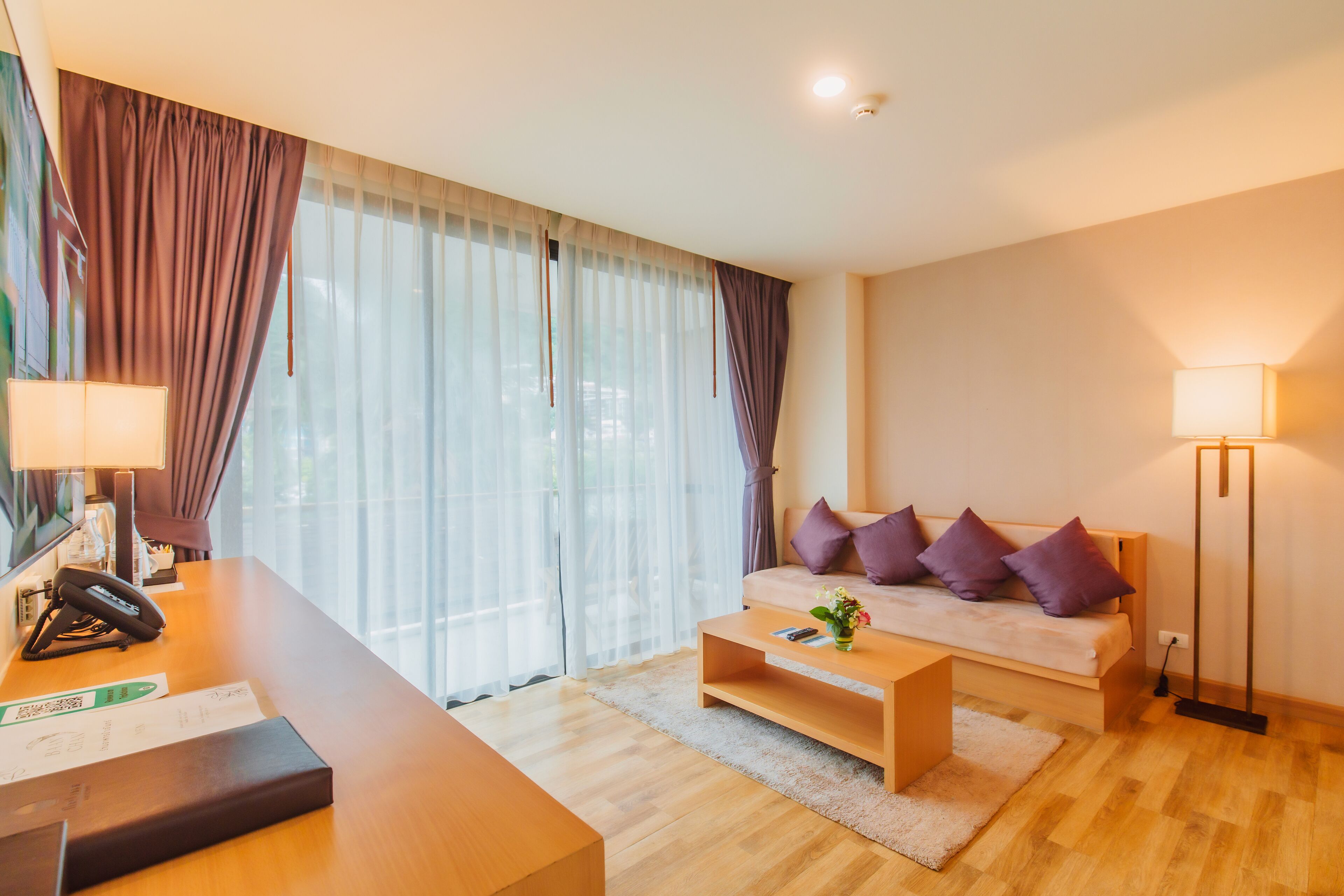 Habitación de hotel con un escritorio de madera, una cama con almohadas moradas, una lámpara de pie y un ventanal con cortinas translúcidas.