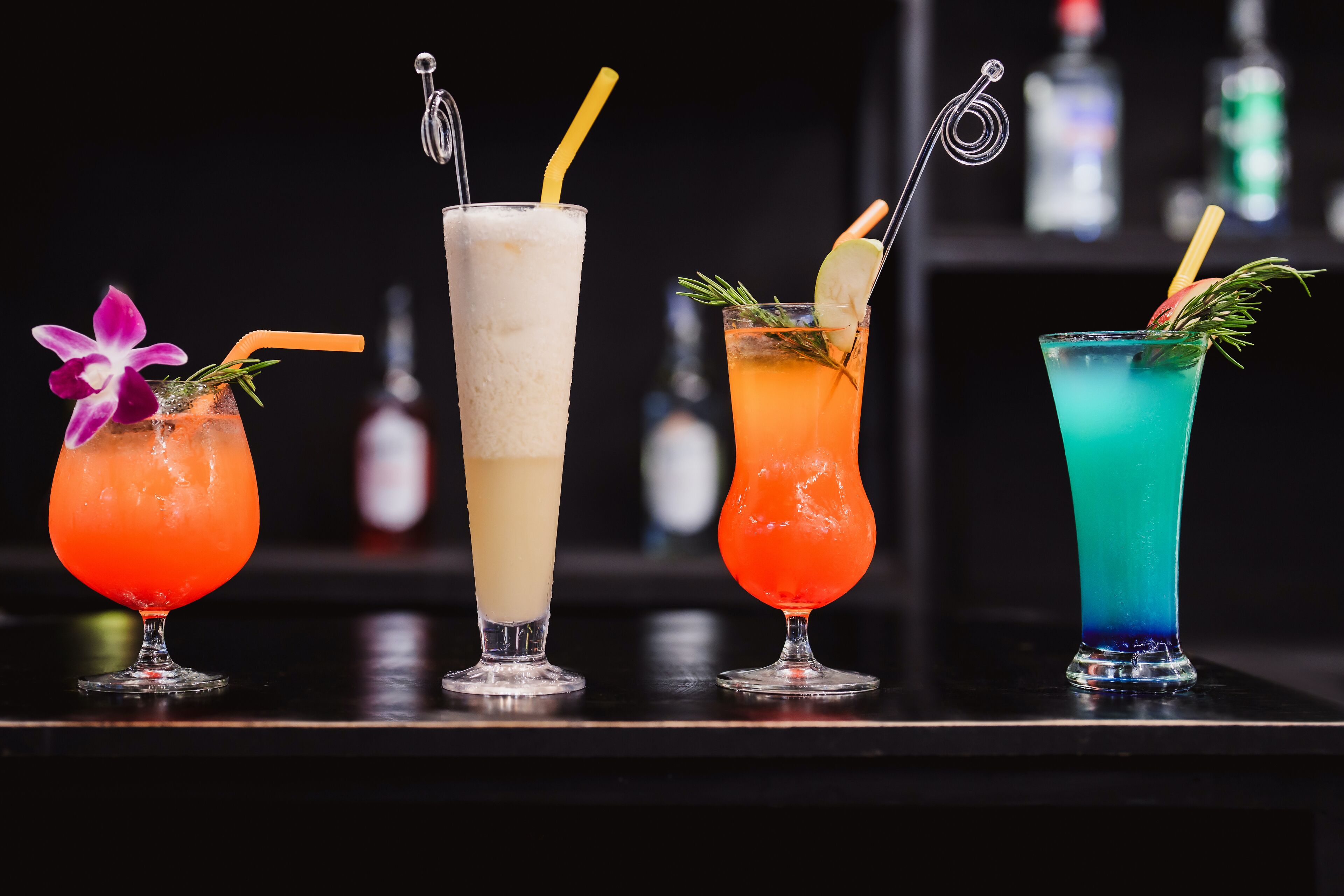 Four cocktails with straws and garnishes on a bar counter.