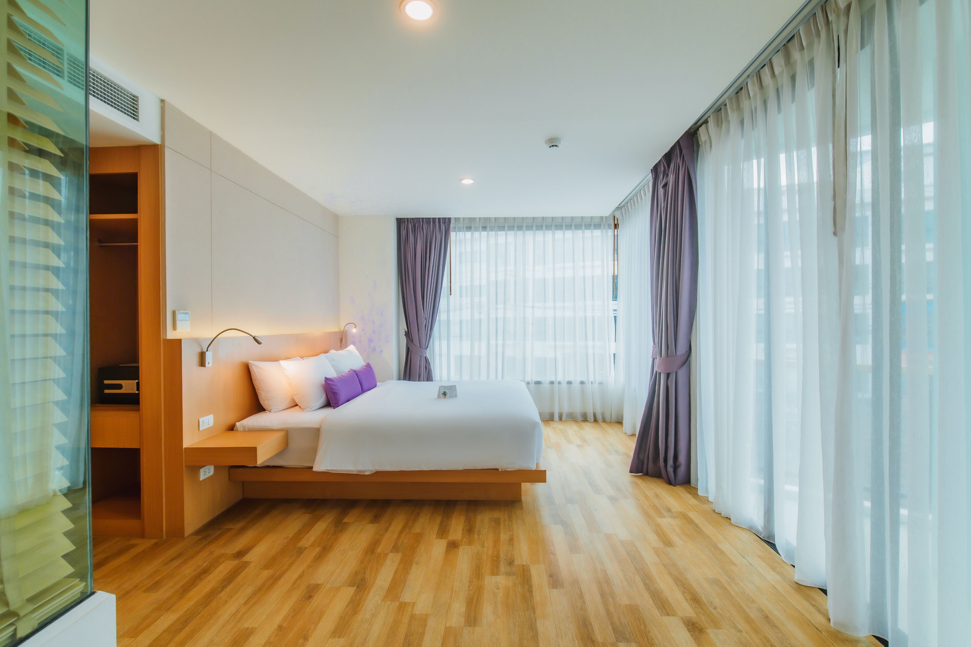 A modern hotel room with a wooden floor, a bed with white bedding, a bedside table, and a large window with sheer curtains.
