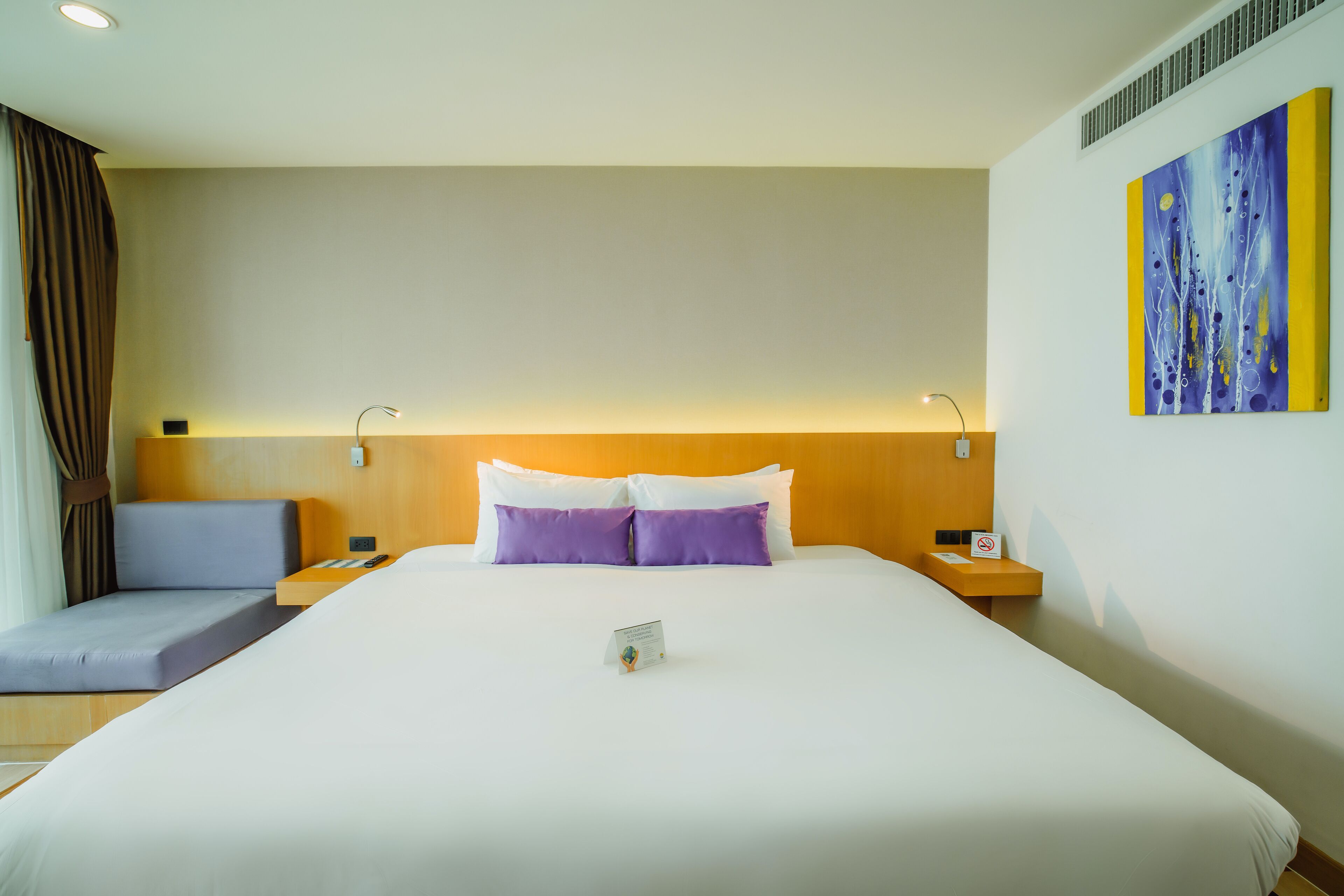 Une chambre d’hôtel avec un grand lit, deux tables de chevet, une petite table, une œuvre d’art accrochée au mur et une fenêtre avec des rideaux.