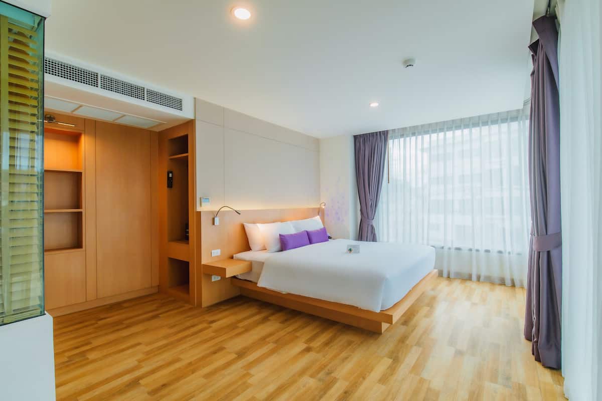 Une chambre d’hôtel moderne avec un plancher en bois, un grand lit avec une literie blanche et violette, une armoire intégrée et une grande fenêtre avec des rideaux.