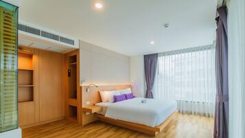 Ein modernes Hotelzimmer mit Holzboden, einem großen Bett mit weißer und violetter Bettwäsche, einem eingebauten Kleiderschrank und einem großen Fenster mit Vorhängen.