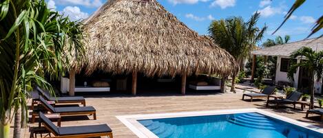 Zona de piscina con sillones de descanso, una estructura de techo de paja y palmeras.