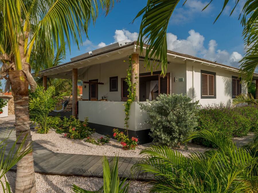 Une maison de plain-pied dotée d’un porche abrité, entourée d’une végétation tropicale et surplombée d’un ciel bleu azur.
