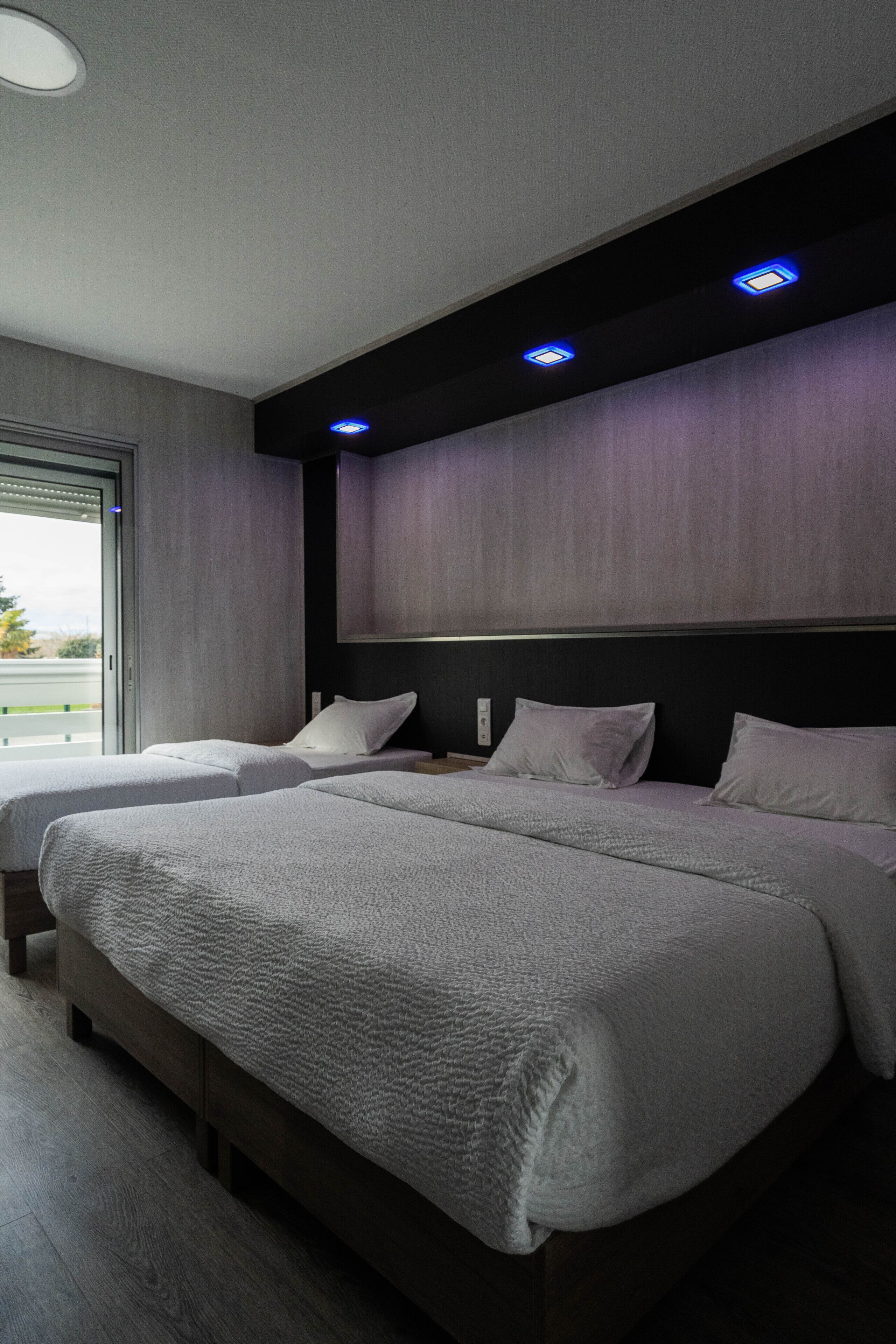Una habitación de hotel moderna con dos camas, un ventanal grande y un techo oscuro con iluminación empotrada.