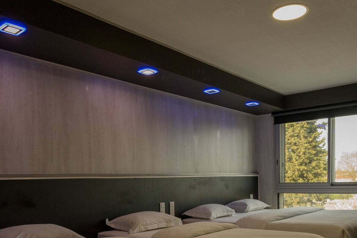 A hotel room with three beds, wooden flooring, and a large window with a view of trees.
