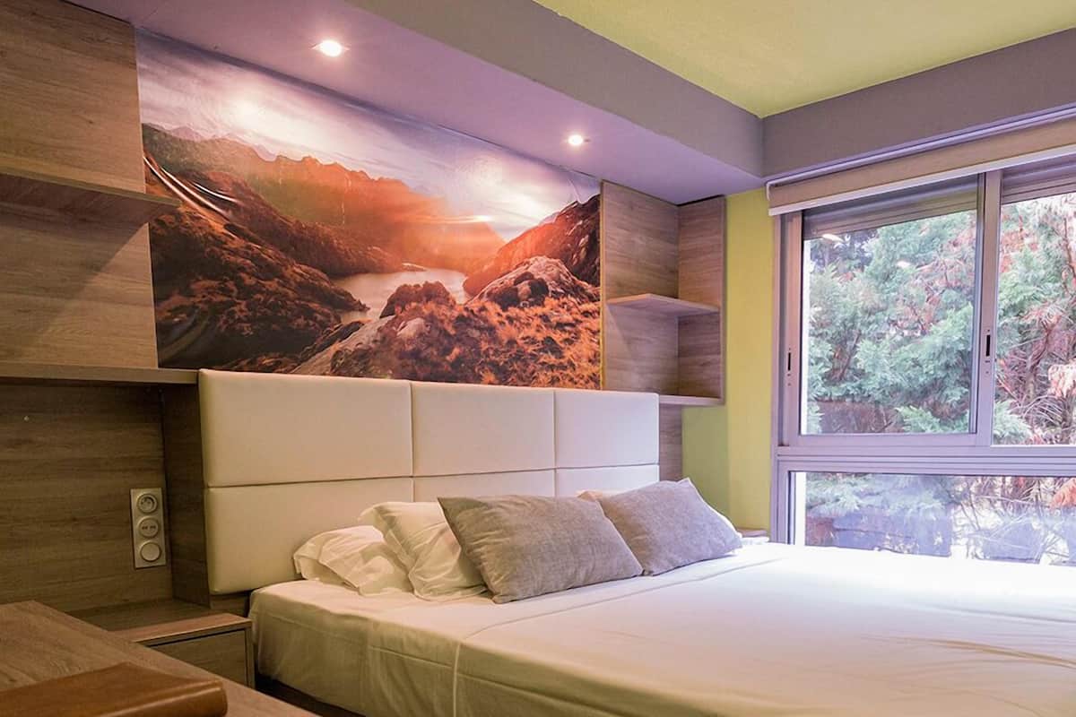 A modern bedroom with a large bed, a nightstand, a bench, and a window with a view of trees.